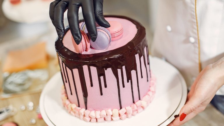 Anonymous female cook in uniform and latex gloves decorating cake with macaroons in light kitchen